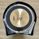 Dvě mosazné činely v černém plastovém kufru, přikryté průhlednou fólií. Na horním činele je viditelné logo Paiste. Mezi činely je umístěna černá tlum…