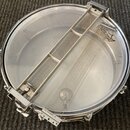 Fotografie zachicuje pohled zevnitř na snare drum (malý buben) s kovovým obručováním. Uvnitř je vidět napnuté pletivo s kovovou lištou, na které je č…
