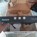 Černý DJ mixér s nápisy Numark a PARTY MIXER LIVE. Zařízení má na přední straně tři kulaté kontrolky s průhledným krytem. Mixér je držen v ruce nad o…