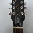 Fotografie zachycuje hlavu elektrické kytary v černé barvě s chromovanými ladičkami. Na horní části je zlatý nápis Gibson, uprostřed nápisy Les Paul…
