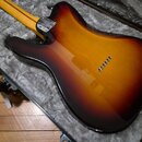 Zadní strana elektrické kytary s tvarem typu Stratocaster v lakování sunburst (přechod od žluté k černé). Nástroj leží v otevřeném kufru s šedým pols…