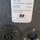 Fotografie detailu bílého typového štítku na černém, strukturovaném povrchu. Na štítku je text: MODEL E122, SER. NO. 12533, IMPEDANCE 8 OHMS, POWER R…