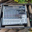 Stříbrný mixážní pult značky behringer s nápisem XENYX QX1832USB umístěný v černé dřevěné bedně. Zařízení obsahuje numerous červené a šedé otočné kno…