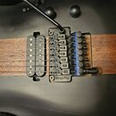 Černá elektrická kytara s hnědým dřevěným pruhem uprostřed těla. Nástroj má dva černé humbuckery a černý kobylník s nápisem FloydRose. Na hrazníku je…