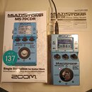 Fotografie zobrazuje modrý efektový pedál Zoom MultiStomp MS-70CDR, jeho originální krabici a uživatelskou příručku. Na pedálu je vidět zapnutý displ…