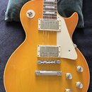 Elektrická kytara s žlutým trupem a hnědým hmatníkem s trapezoidovými vložkami. Nástroj má dva humbuckery s kovovými kryty, bílý plechtrum a čtyři zl…