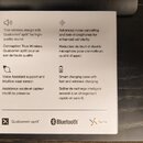 Fotografie zachycuje bílý informativní leták s anglickým a francouzským textem popisujícím funkce bezdrátových sluchátek. V dolní části jsou loga Qua…