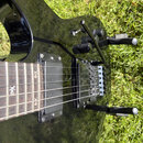 Černá elektrická kytara s dvěma humbuckery a tremolo systémem. Na hrazdici jsou viditelné vložky s bílým symbolem připomínajícím stylizované písmeno…