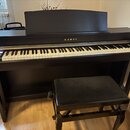 Černý digitální piano s bílými a černými klávesami. Na přední straně nad klaviaturou je zlatý nápis KAWAI. Součástí je černá pianová stolička s polst…