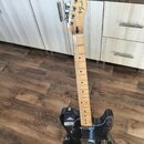 Černá elektrická kytara s jasným dřevěným hnedým krčkem a hmatníkem. Na hlavě je viditelné logo Fender® a nápis TELECASTER®. Povrch těla vykazuje dro…