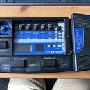 Černý kytarový procesor s modrým ovládacím panelem. Na panelu je nápis MODELING SIGNAL PROCESSOR AX100G a vpravo na boku přístroje logo TONEWORKS KOR…