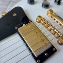 Detail bílé elektrické kytary se zlatým hardwarem. Na snímku je vidět jeden zlatý krytý snímač s šestnácti šroubky, černý plastový pickguard a zlatý…