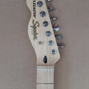 Hlava elektrické kytary z přírodního světlého dřeva s šesti kovovými ladičkami. Na hlavě je černý nápis TELECASTER® a zlaté logo Squier s menším nápi…