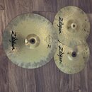 Tři mosazné činely v různých velikostech položené na hnědém laminátovém povrchu. Každý činel nese černý nápis Zildjian a logo PLANET Z. Povrchy činel…