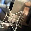 Kondenzátorový mikrofon v béžové barvě s kovovou mřížkou, umístěný v bílém pavoukovém držáku. Na těle mikrofonu je viditelné červené logo s ptačím sy…