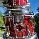 Detail červeného bubnu s glitrovým povrchem a chromovaným kováním. Na chromovaném držáku je vidět logo Pearl a nápis Optima. Povrch nástroje i kovové…