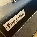 Detail kytarového komba s černým texturovaným povrchem a šedou přední síťovkou. Na horní části je bílá obdélníková tabulky s hnědým nápisem FRIEDMAN.…