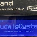 Detailní záběr na displej a horní část černého modulu elektronických bicích. Na šasi je bílý nápis Roland, DRUM SOUND MODULE TD-30 a logo SuperNATURA…
