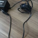 Černý napájecí adaptér s připojeným kabelem. Na boku adaptéru je viditelný bílý nápis „UPPLY“, pod ním „00mA“ a dále „EFFECTS-“. Povrch adaptéru je t…