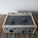Šedý audio procesor v dřevěném rámu. Na horním panelu je logo UA, nápisy INPUT a OUTPUT s LED indikátory, přepínač s označením VOC, GTR, FAST, OFF a…