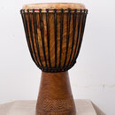 Dřevěný buben typu djembe s přirozenou hnědou barvou dřeva a viditnou strukturou zrn. Membrána je natažená pomocí černých provazů, které jsou v horní…