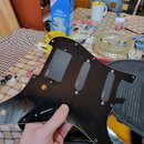 Černý plastový pickguard pro elektrickou kytaru s třemi výřezy pro snímače. Na každém z nich je umístěn černý kryt s bílým nápisem ETIG. Povrch pickg…