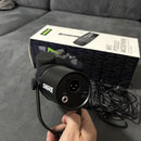 Černý dynamický mikrofon značky SHURE v držáku. Na zadní straně jsou vidět konektory XLR, USB-C a vstup pro sluchátka s ikonou. V pozadí je bíločerná…