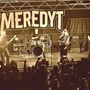 Fotografie zachycuje kapelu během koncertu na pódiu před publikem. V pozadí visí černý banner s bílým nápisem MEREDYT. Na pódiu jsou vidět hudební ná…