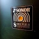 Detail černé čtvercové zásuvky s bílými nápisy a grafickými prvky v podobě koncentrických kruhů. V horní části je logo a nápis SONOR, ve spodní části…