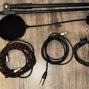 Sada příslušenství rozložená na dřevěné podlaze. Obsahuje černý mikrofonní stojan, černý popfiltr a tři audio kabely (jeden s červeným provázekem, dv…