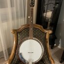 Banjo s bílou membránou, hnědým dřevěným rímem a krčkem. Na hlavě nástroje je viditelné logo, které není čitelné. Nástroj je opřen o zdobenou židli s…