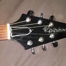 Černá hlava elektrické kytary s nápisem Epiphone. Jsou na ní vidět šest ladicích kolíků a hřif s prázdnými políčky. Povrch je lesklý, v místě nápisu…