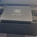 Černá papírová krabice s vnitřní pěnovou výplní. Na víku je bílý nápis AMP TOP BOX a logo ox v obdélníkovém rámečku. Povrch krabice vykazuje mírné po…