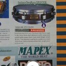 Fotografie zachycuje výstřižek z katalogu s malým bubnem (piccolo snare) značky TAMA. Nástroj má tmavý korpus s kovovými obručemi a uprostřed je vidi…