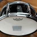 Černý buben s chromovaným obručováním a mechanismy. Na bílém koženu je vidět logo REMO a nápis SILENTSTROKE™ MADE IN U.S.A. Povrch nástroje je glossy…