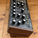 Černý analogový syntetizátor Moog MOTHER-32 v dřevěném podstavci. Na horním panelu jsou umístěny početné otočné knofíky, přepínače a patchovací konek…