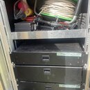 Fotografie zachycuje otevřený transportní case s hudebním vybavením. V horní části je zelená cívka s namotaným bílým kabelem, černý box a různé konek…