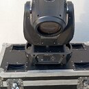 Černý pohyblivý reflektor typu moving head umístěný na černém transportním kufru s hliníkovými hranami a kovovými zámky. Na horní části přístroje je…