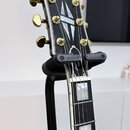 Černá elektrická kytara s bílým lemováním, umístěná na černém stojanu značky HERCULES CUSTOM. Na hlavě nástroje je bílé logo Epiphone a vložka s geom…