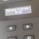 Černý elektronický modul s bílým logem Roland. Na displeji je nápis Roland V-Drums TD-07. Pod displejem se nacházejí čtyři ovládací tlačítka s ikonam…