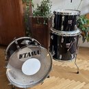 Černá bubnová sada tvořená basovým bubnem a dvěma tomami. Na basovém bubnu je viditelné logo TAMA a na spodním obruči nápis TAMA. Oba tomové bubny ma…