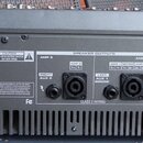 Zadní strana černého mixážního pultu s nápisem SAMSON TXM20 1000W STEREO POWERED MIXER. Jsou zde vidět dva výstupy SPEAKER OUTPUTS s popisky AMP 2, R…