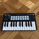 Černý kompaktní MIDI kontrolér s klávesnicí a bílými padovými tlačítky. Na zařízení jsou nápisy novation, LAUNCHKEY MINI a IN CONTROL. Povrch je v do…