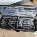 Otevřený černý kufr na hudební nástroj s vnitřním polstrováním z šedého plyšového materiálu. V dolní části jsou vidět tři oddělené přihrádky pro ulož…