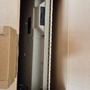 V kartonové krabici je umístěn bílý elektronický přístroj s vertikálním uspořádáním. Na přední straně jsou vidět dva tmavé obdélníkové prvky, pravděp…