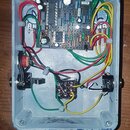 Interiér světle modré kovové krabice s elektronickým obvodem na desce. Na desce jsou viditelné součástky a nápisy 47nJ100 a 202000. V dolní části jso…