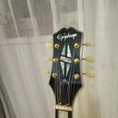 Hlava elektrické kytary v černé barvě s bílým lemováním. V horní části je bílý nápis Epiphone a uprostřed se nachází vložka ve tvaru kříže z bílého m…