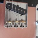 Detail elektrické kytary v růžové barvě s kovovým mostem a černým pickguardem. Na kovovém krytu je vyryto „FENDER PAT. PEND.“. Most má zlaté sedla a…