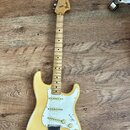 Světle žlutá elektrická kytara s bílým pickguardem a hnedým hmatníkem z jednoho kusu dřeva. Na hlavě je černý nápis Fender STRATOCASTER. Nástroj vyka…