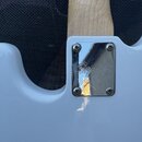 Detail zadní strany bílé elektrické kytary v místě spoje krku s tělem. Na chromovaném krytu krku je vyryt nápis Squier®. Kryt je připevněn čtyřmi šro…